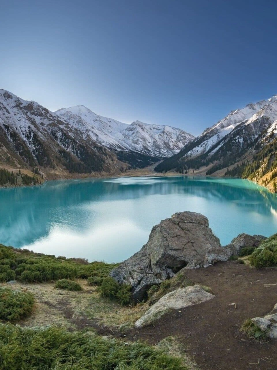 Panoramic view of Big Almaty Lake surrounded by snowy peaks and rocky slopes.
