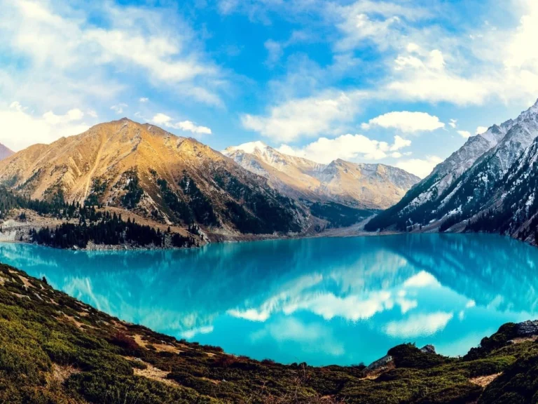 Wide panoramic shot of Big Almaty Lake with bright turquoise water and surrounding mountain ridges.