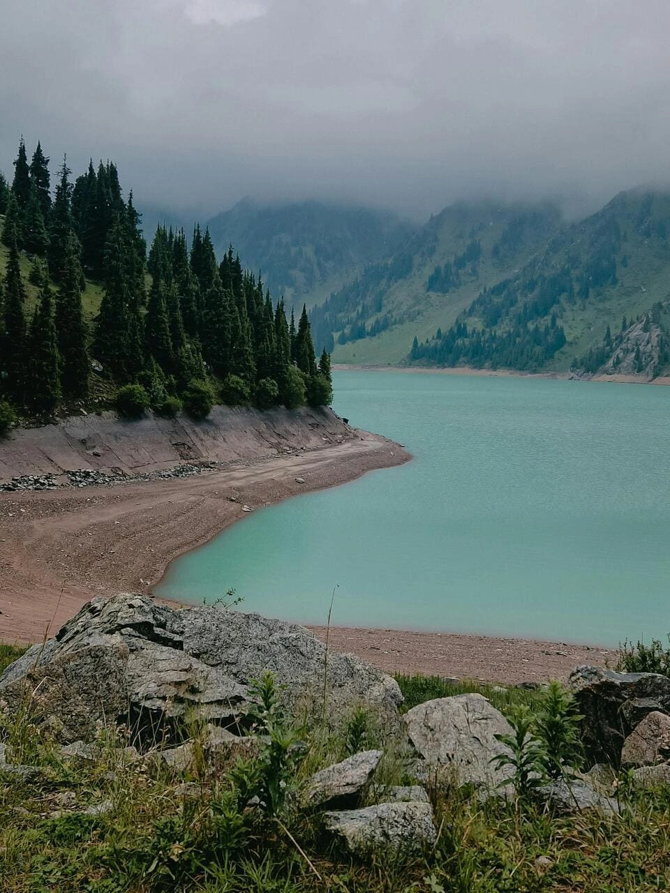 Foggy morning at Big Almaty Lake with pine forests and soft low-hanging clouds.