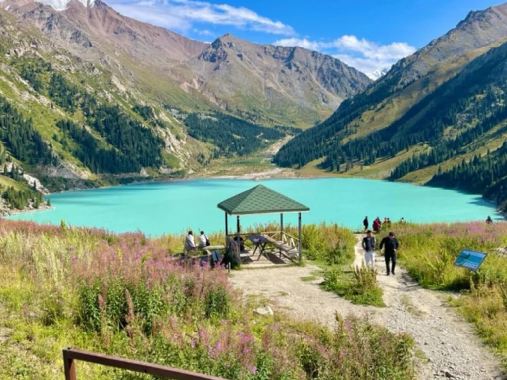 View of Big Almaty Lake with turquoise water, mountain peaks, and a wooden pavilion in the foreground.
